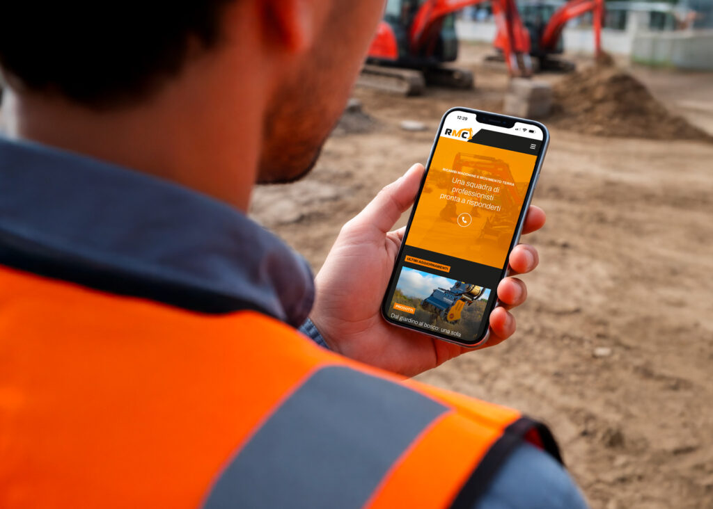 Operatore in cantiere consulta il nuovo sito web di RMC su smartphone con escavatori sullo sfondo.