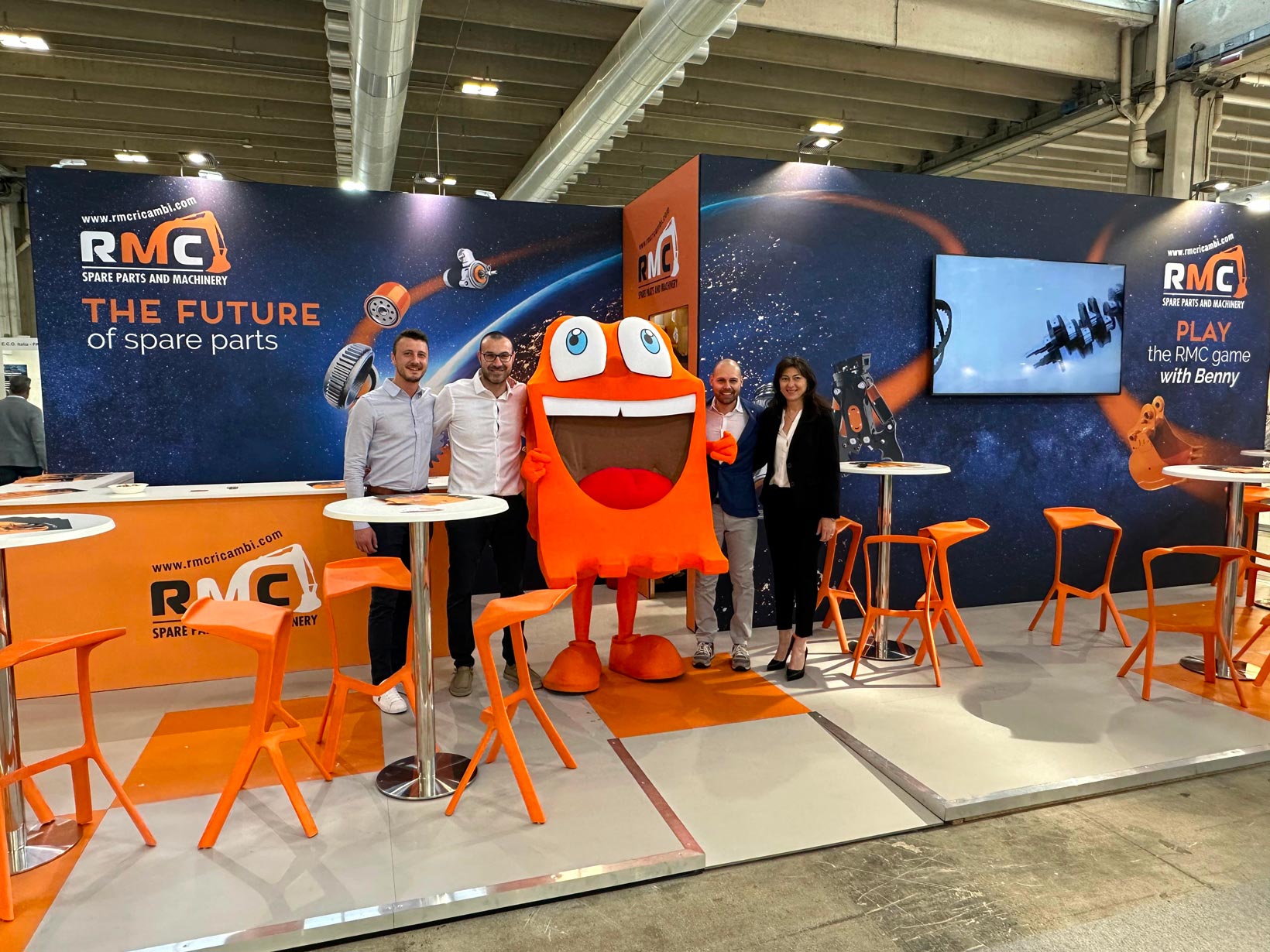 Team RMC allo stand fieristico aziendale insieme alla mascotte Benny durante un evento di settore.