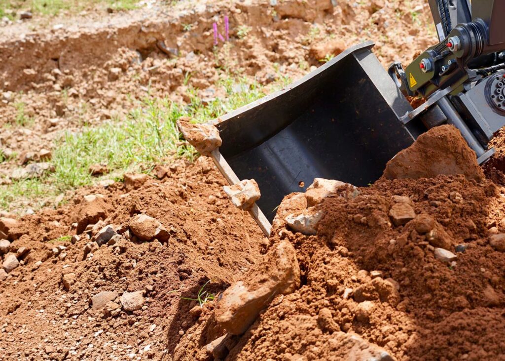 Benna di escavatore con denti durante operazione di scavo su terreno misto.