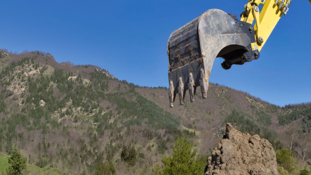 Benna di escavatore con denti in primo piano durante attività di scavo in ambiente naturale.