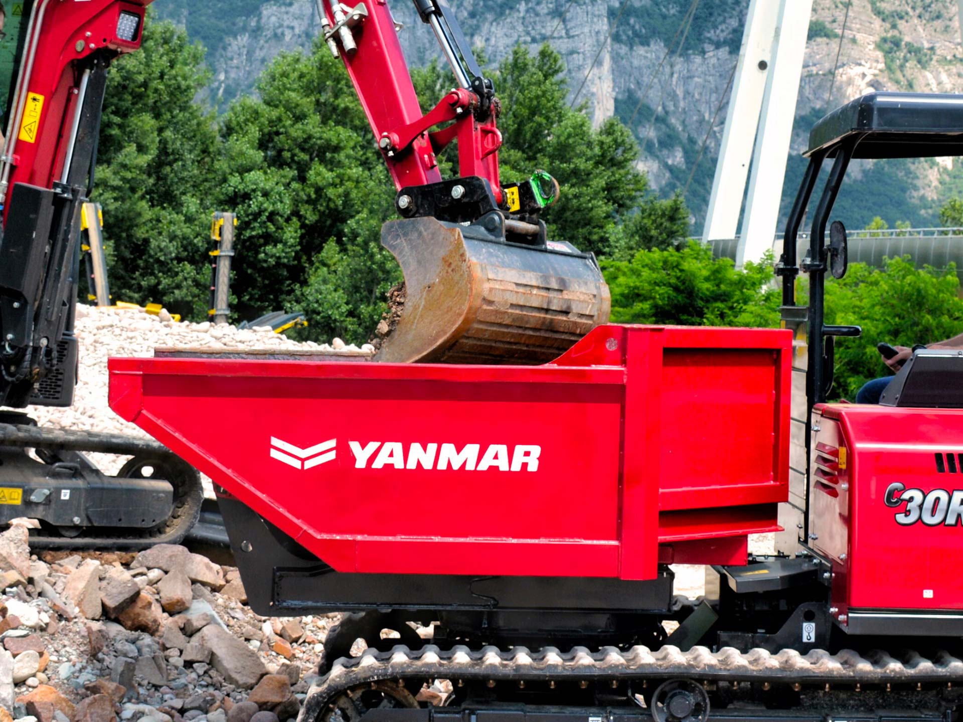 Escavatore carica materiale nella vasca di un dumper cingolato Yanmar C30R durante le operazioni di movimentazione in cantiere.