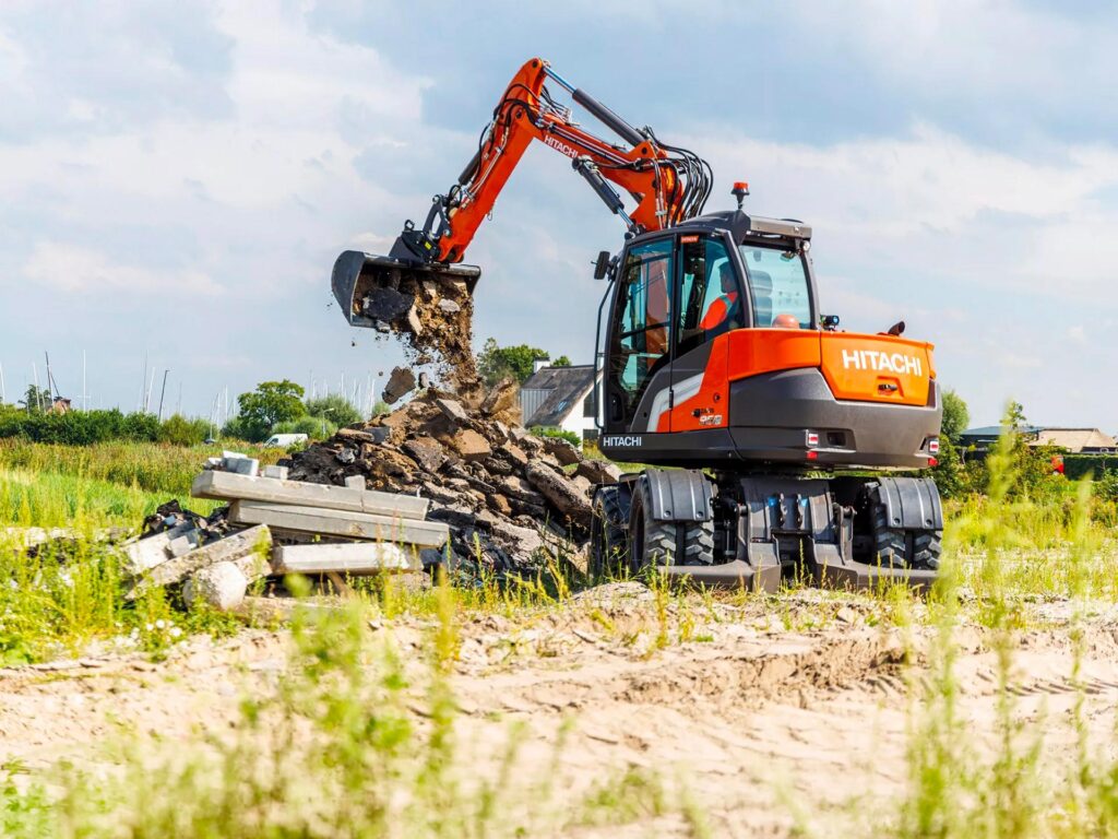 Escavatore gommato Hitachi al lavoro in un cantiere stradale durante operazioni di movimentazione di materiale da demolizione.