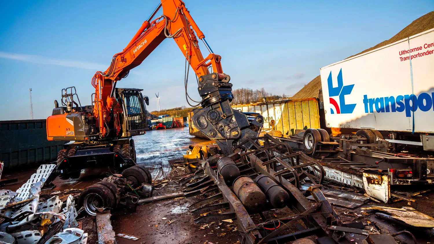 Escavatore gommato Hitachi con pinza idraulica durante operazioni di demolizione e movimentazione di rottami metallici in un impianto di recupero.