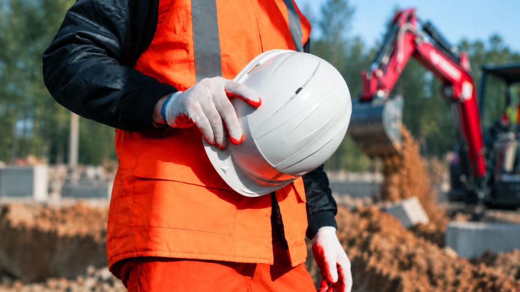 Operatore con giubbotto ad alta visibilità e casco di sicurezza in un cantiere con escavatore sullo sfondo.